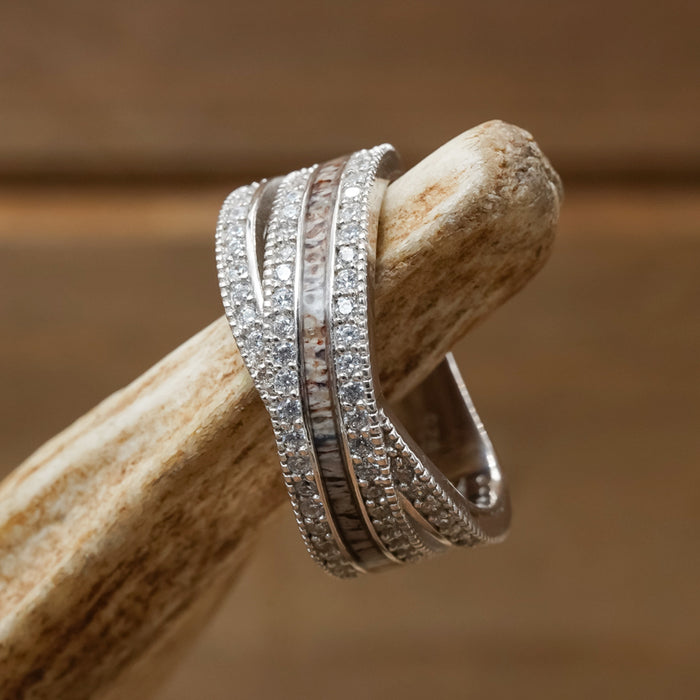 The Chelly, a multi-band sterling silver ring featuring small, sparkling stones and patterned inlays, is shown draped over a piece of antler against a softly blurred brown background.