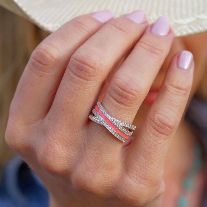 A hand with light pink manicured nails showcases The Chelly—a stylish sterling silver ring featuring intertwined silver and pink bands—while holding a light-colored hat that partially obscures the face.