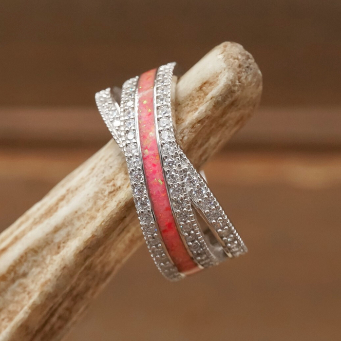 Introducing The Chelly: a stunning women's band crafted in sterling silver, featuring wavy bands adorned with small clear stones and a central strip of pink opal, beautifully presented on a piece of light-colored wood.