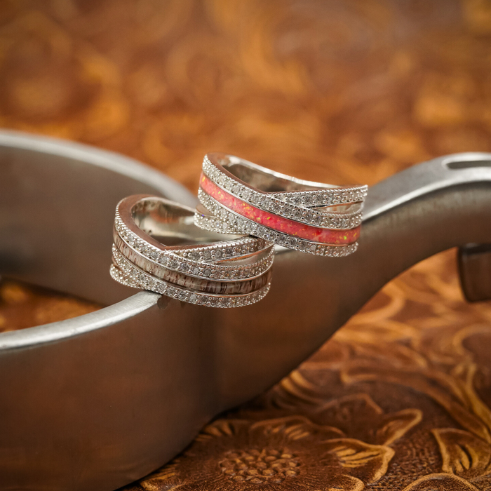 Two sterling silver rings from The Chelly collection, each with sparkling stones and colorful inlays—one highlighted by a vivid red stripe, the other adorned with a distinctive brown antler ring inlay—rest on a metallic surface against a patterned brown background.