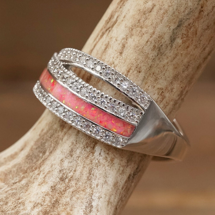 The Chelly is a sterling silver ring featuring two rows of small clear stones and a center band of pink opal inlay, elegantly displayed on a beige, textured surface. This sophisticated women's band effortlessly elevates any ensemble.