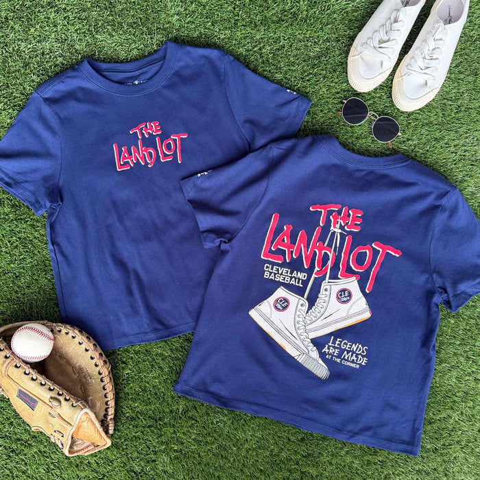 Two blue t-shirts with 'The Land Lot' design on a grass background with sports equipment.