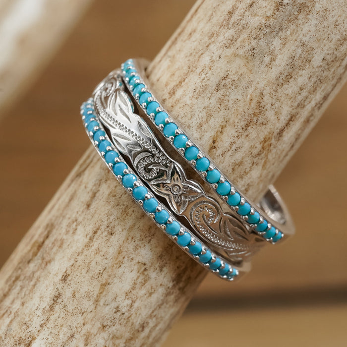 The Paria - Promo! features two stacked sterling silver rings—one engraved with a delicate floral pattern, the other adorned with small turquoise stones—displayed on light-colored wood for a timeless stacking set.
