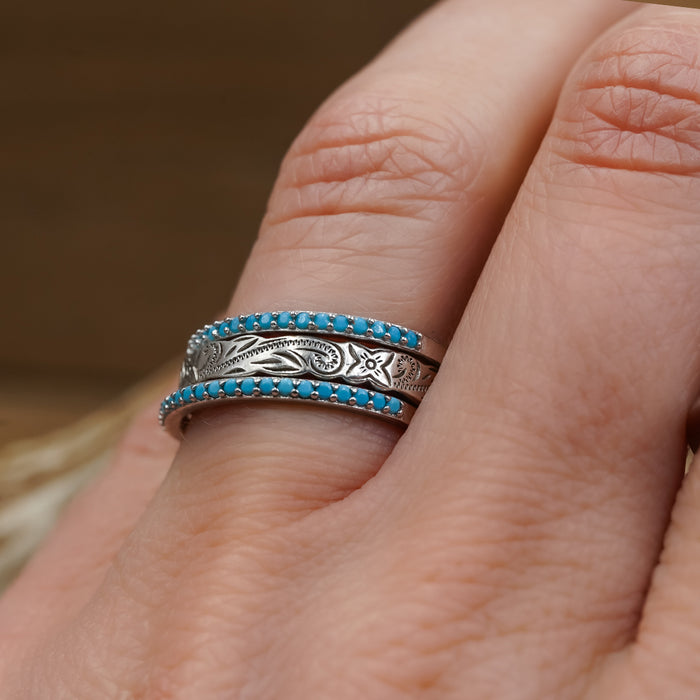 A hand wearing The Paria, a sterling silver ring engraved with floral patterns, flanked by two thin stacking rings set with small turquoise stones.