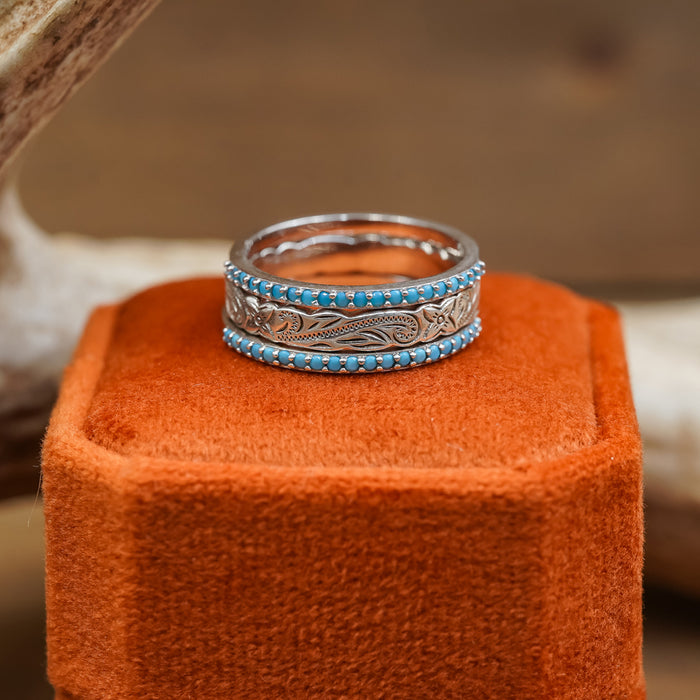 The Paria, a sterling silver ring featuring intricate engraved patterns and a border of small turquoise stones, sits atop an orange velvet display stand. Perfect for wearing solo or stacking with other rings, this piece brings artistic flair to any jewelry collection.