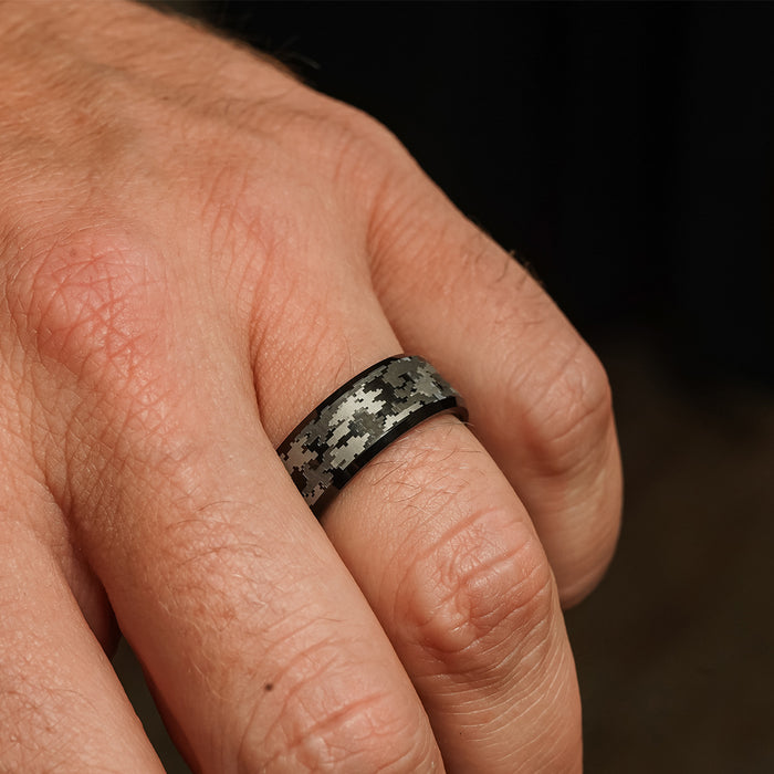A close-up shot of a hand wearing The Sarge, a black tungsten carbide ring featuring a gray camo pattern, on the middle finger with an out-of-focus background.