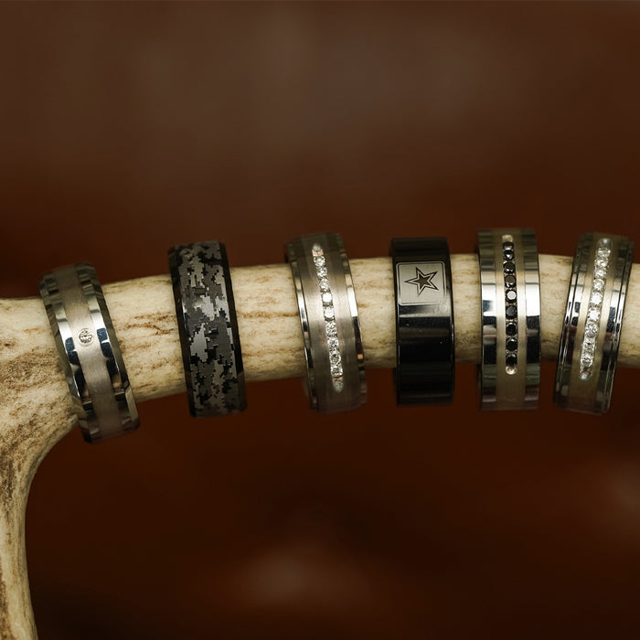 Six metallic rings with various designs, including gemstones, intricate patterns, and a star symbol, are displayed on a piece of antler against a brown background. Featuring "The Sarge" ring, this distinctive collection showcases the versatility of men’s fashion rings—with unique touches such as the rugged tungsten carbide construction of The Sarge and bold digital camo styles—making them perfect for modern trendsetters.