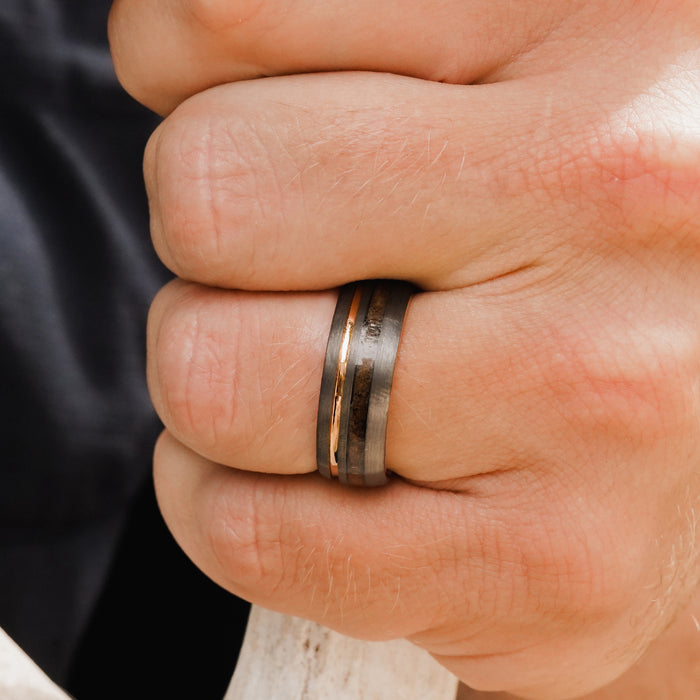 A close-up of a hand wearing The Tikal, a men’s 8mm tungsten ring in gun metal grey featuring a thin gold stripe and a brushed finish, set against a blurred background.