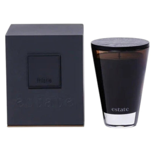 The ZODAX Thistle Estate Candle (10.5 oz) in black is displayed in a glass holder beside its matching box, both reflecting the fresh California coast fragrance.