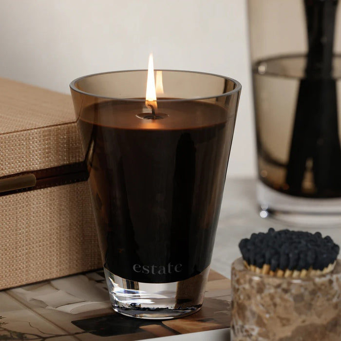 The ZODAX Thistle Estate Candle (10.5 oz) in black is displayed in a glass holder beside its matching box, both reflecting the fresh California coast fragrance.