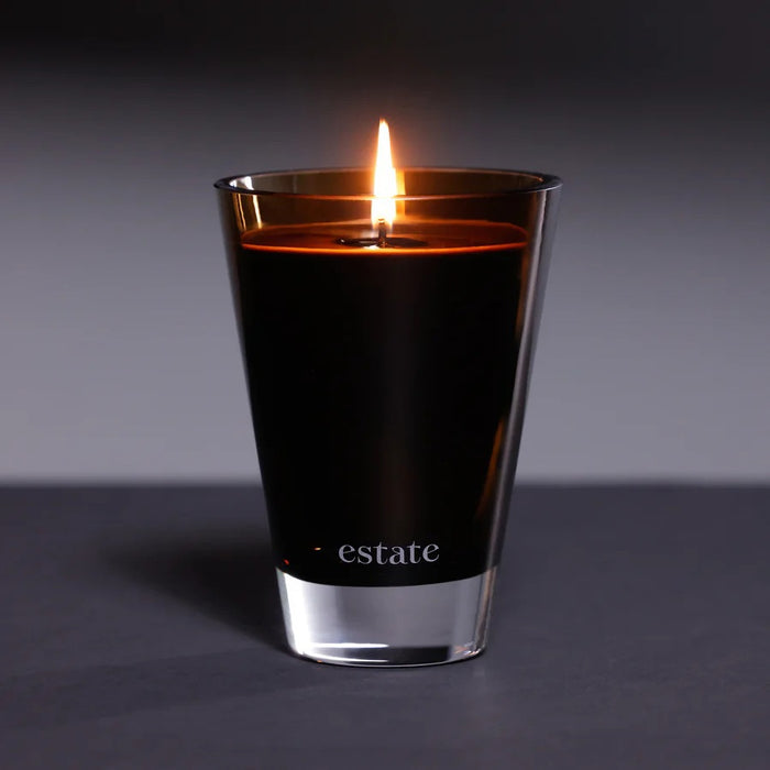 The ZODAX Thistle Estate Candle (10.5 oz) in black is displayed in a glass holder beside its matching box, both reflecting the fresh California coast fragrance.