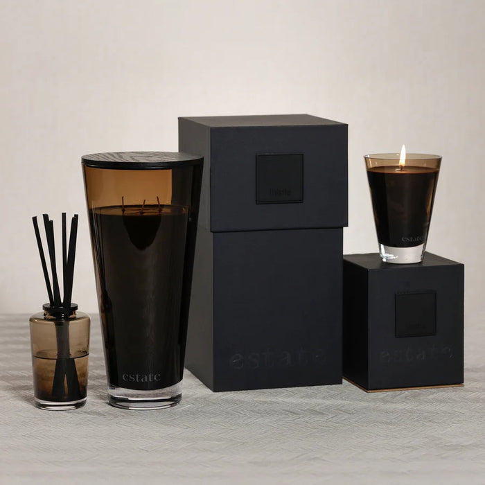 The ZODAX Thistle Estate Candle (10.5 oz) in black is displayed in a glass holder beside its matching box, both reflecting the fresh California coast fragrance.