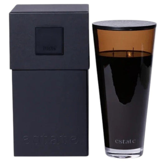 A collection of ZODAX luxury scented candles, including the 57 OZ THISTLE ESTATE CANDLE in black and smoky brown, is displayed on a white textured surface against a plain light background.