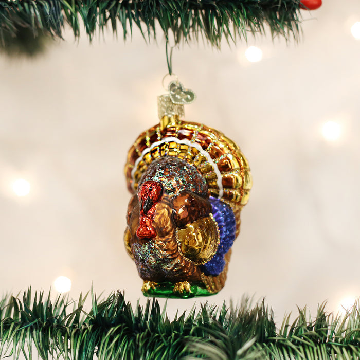 A colorful glass turkey ornament hangs from a green Christmas tree branch, decorated with gold, red, blue, and brown accents. The background is softly blurred with white lights.