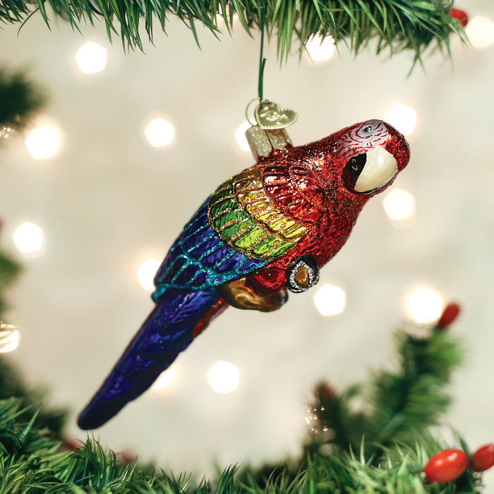 A colorful parrot ornament with red, green, blue, and yellow feathers hangs from a Christmas tree branch, with blurred white lights in the background.
