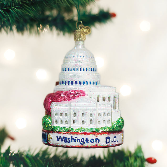 A Christmas tree ornament shaped like the U.S. Capitol building with Washington D.C. written on the base hangs on a green tree branch. The ornament is decorated with white, blue, red, and green accents.