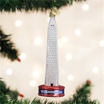 A Christmas tree ornament shaped like the Washington Monument hangs among green branches, with the word Washington at the base and blurred white lights in the background.
