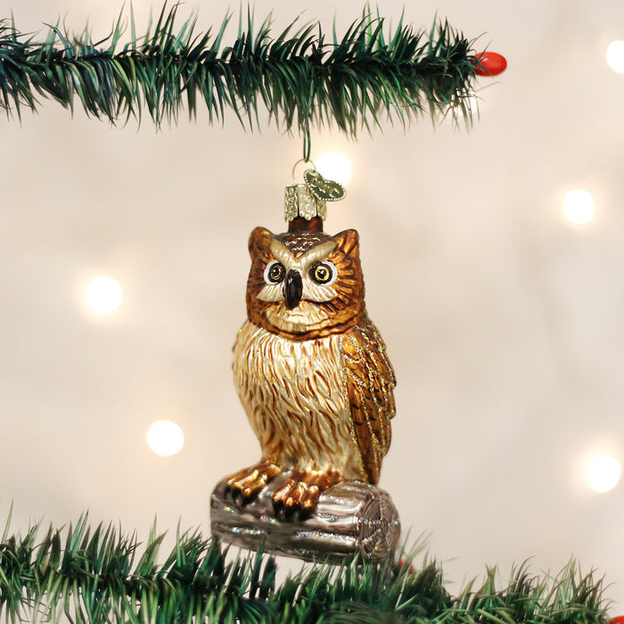 A shiny owl-shaped ornament hangs from a green Christmas tree branch, with blurred white lights in the background. The ornament is gold and brown, perched on a small log.