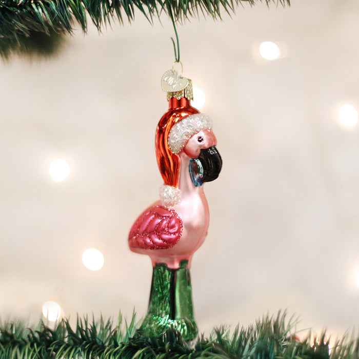 A pink flamingo Christmas ornament wearing a red Santa hat and a black face mask hangs from a green Christmas tree branch, with blurred white lights in the background.